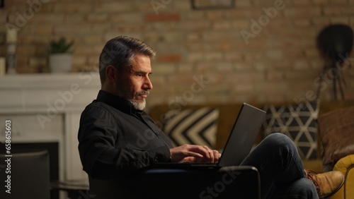 Mid adult businessman with laptop computer in home office, banking online, remote working. Portrait of older gray hair bearded man. Entrepreneur managing business on internet.