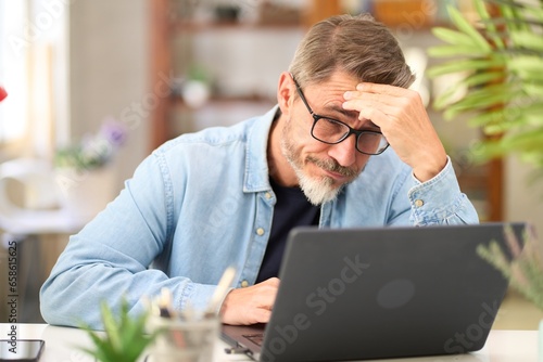 Casual mid adult man with laptop computer at desk in home office, having problem, troubled. Portrait of older gray haired bearded guy thinking. Businessman managing business on internet.  