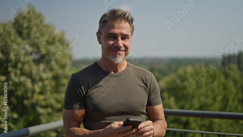 Portrait of happy casual older bearded man outdoor, smiling, Mid adult, mature age guy standing on balcony or terrace, using mobile phone.