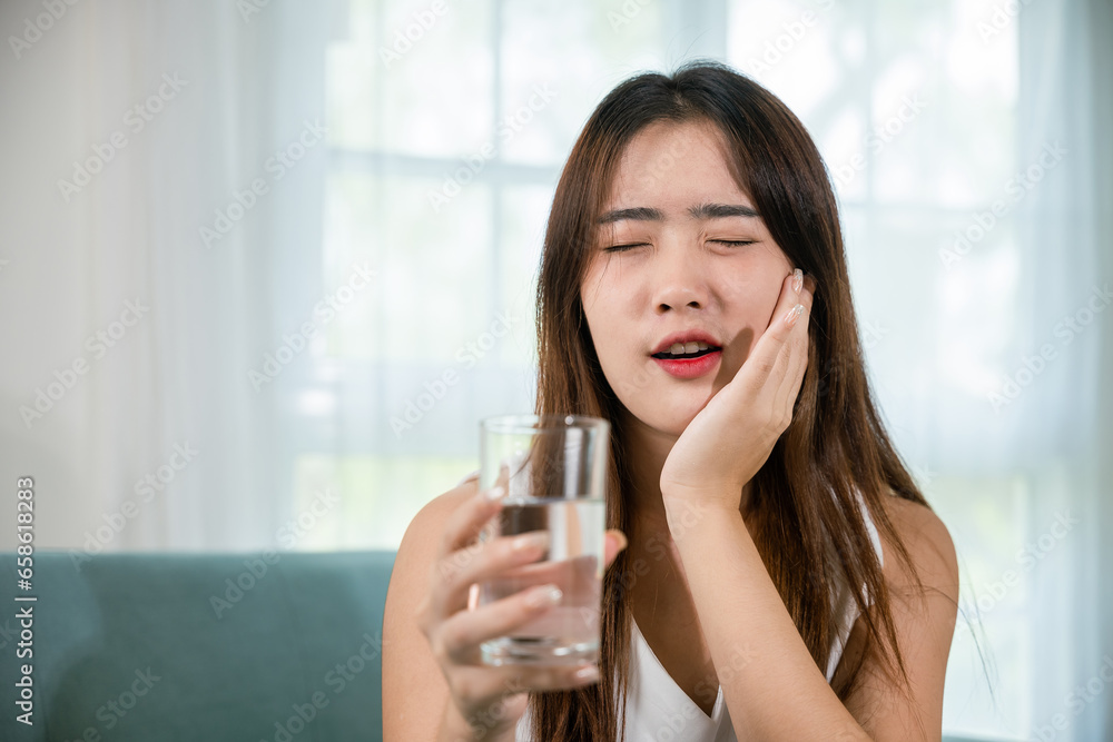 Beautiful female toothache and dental problems touching cheek feeling pain after drinking cold water, Asian young woman with sensitive teeth holding glass of cold water at home in living room, pain Beautiful female toothache and dental problems touching cheek feeling pain after drinking cold water, Asian young woman with sensitive teeth holding glass of cold water at home in living room, pain