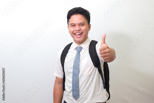 Indonesian senior high school student wearing white shirt uniform with gray tie smiling and looking at camera, making thumbs up hand gesture. Isolated image on white background photo
