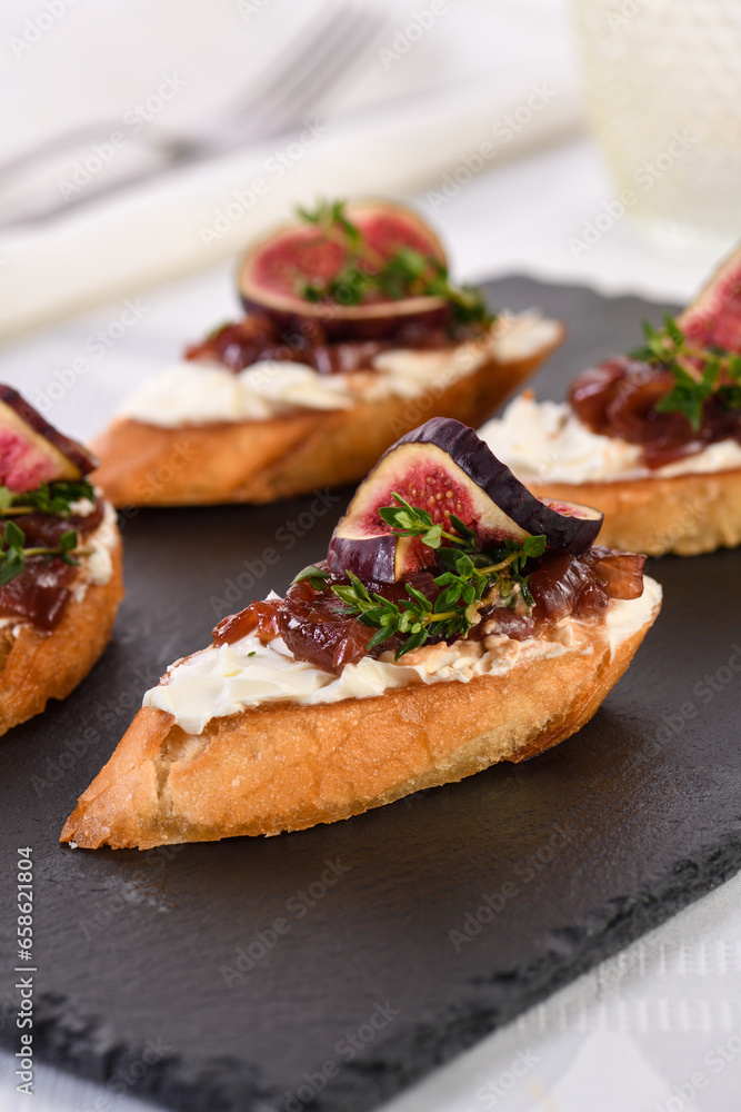 Canape or crostini with toasted baguette, cream cheese, onion jam, figs and fresh thyme on a slate board. Perfect as an aperitif for a holiday or party.