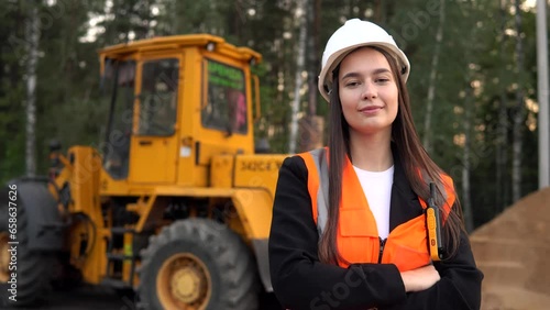 A young woman mine worker is standing in front of a large dump truck or grader. Looking at camera.