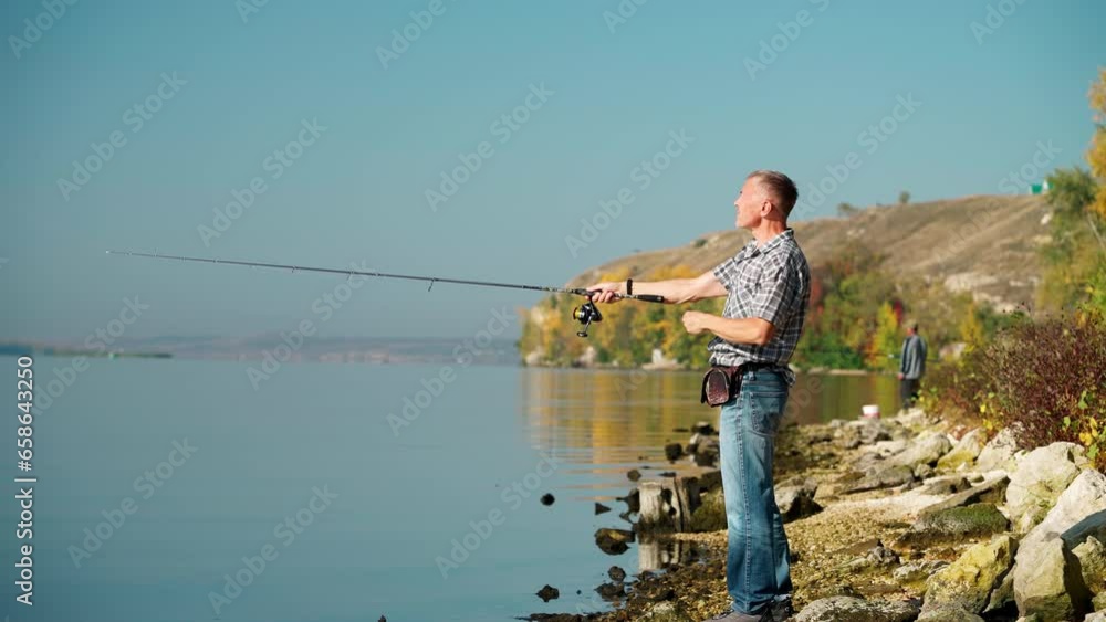 A mature male fisherman in casual clothes casts a lure with a spinning rod while fishing on the river bank. Profile view of an adult man spinning fishing in the weekends. Copy space. A mature male fisherman in casual clothes casts a lure with a spinning rod while fishing on the river bank. Profile view of an adult man spinning fishing in the weekends. Copy space.