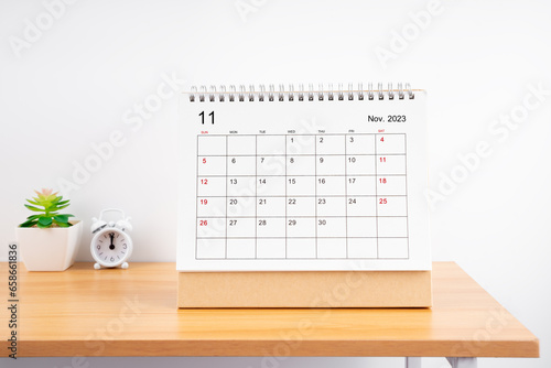 The November Monthly desk calendar for 2023 year and alarm clock on wooden table.