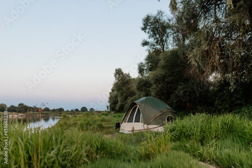 Beautiful summer landscape by the river in the early morning. The tent stands among green grass and trees, on the shore of a lake. Camping. Meeting the dawn by the river.