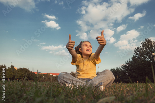 happy smiling cheerful girl 6 years old sits on grass in park and shows gesture perfectly in summer day