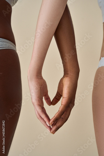 Two diverse women wearing underwear touching hands making heart sign together standing on beige background. Diversity body positive beauty, different ethnicities female help unity connection. Vertical