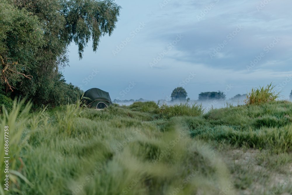Beautiful summer landscape by the river in the early foggy morning. The tent stands among green grass and trees, on the shore of a lake. Camping. Watching dawn near a river covered in thick fog Beautiful summer landscape by the river in the early foggy morning. The tent stands among green grass and trees, on the shore of a lake. Camping. Watching dawn near a river covered in thick fog