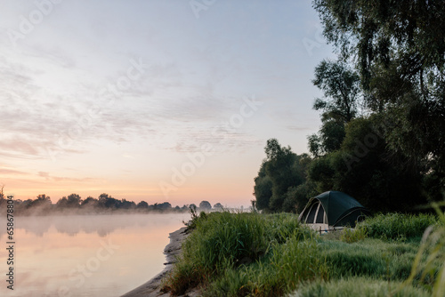 Beautiful summer landscape by the river in the early foggy morning. The tent stands among green grass and trees  on the shore of a lake. Camping. Watching dawn near a river covered in thick fog