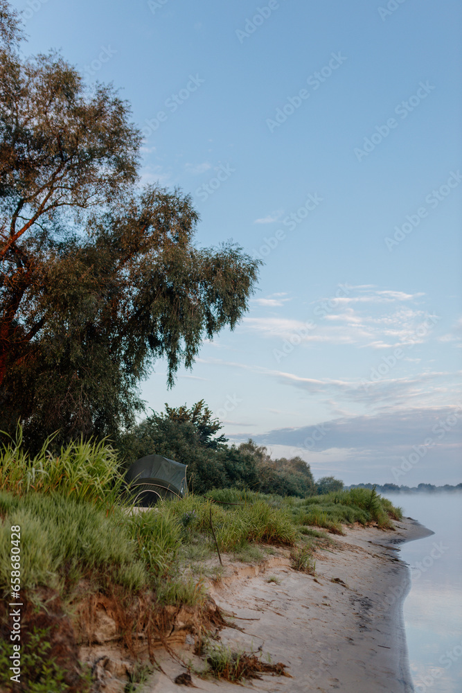 Beautiful summer landscape by the river in the early foggy morning. The tent stands among green grass and trees, on the shore of a lake. Camping. Watching dawn near a river covered in thick fog Beautiful summer landscape by the river in the early foggy morning. The tent stands among green grass and trees, on the shore of a lake. Camping. Watching dawn near a river covered in thick fog