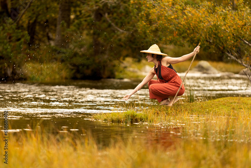 Fototapeta Naklejka Na Ścianę i Meble -  Woman with straw hat in middle of forest with river in background trees and nature warm colors, meditating in nature, traveling through east