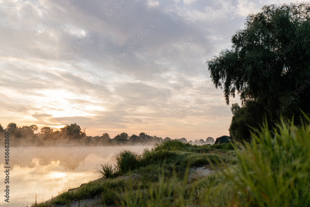 Beautiful summer landscape by the river in the early foggy morning. The tent stands among green grass and trees, on the shore of a lake. Camping. Watching dawn near a river covered in thick fog Beautiful summer landscape by the river in the early foggy morning. The tent stands among green grass and trees, on the shore of a lake. Camping. Watching dawn near a river covered in thick fog