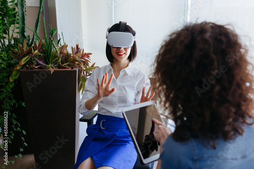 sitting smiling asian woman wearing viewer exploring virtual reality with her left handed colleague working with tablet