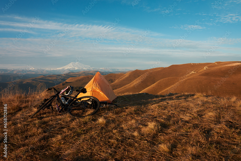 A cyclist's overnight stay in the mountains. Yellow tourist tent, next to a bicycle on the background of mountainous terrain with snow-covered volcanoes in the distance in the early morning.