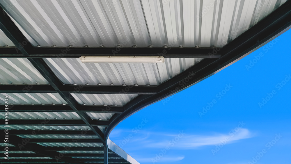 Modern corrugated metal curve roof of covered walkway with black steel roof beam structure against blue sky background, view from below 