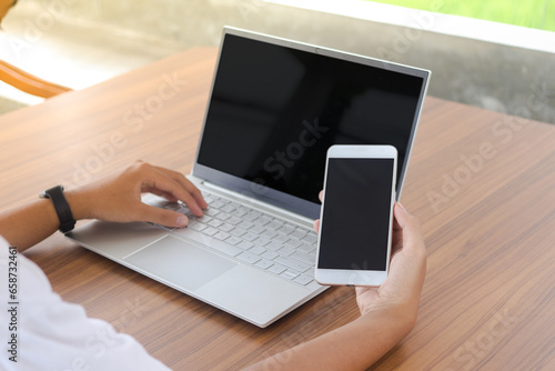 Close up of businessman hand touching blank screen of mobile phone with laptop beside on the table. Display of smartphone and laptop mock up