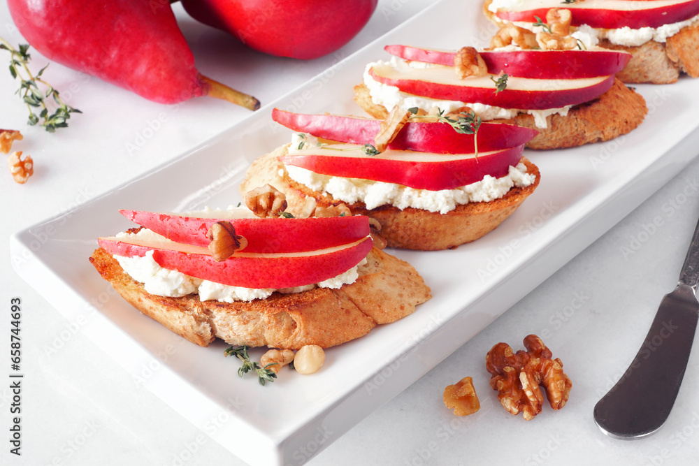 Crostini appetizers with red pears, whipped feta cheese and walnuts. Close up table scene against a white background. Party food concept.