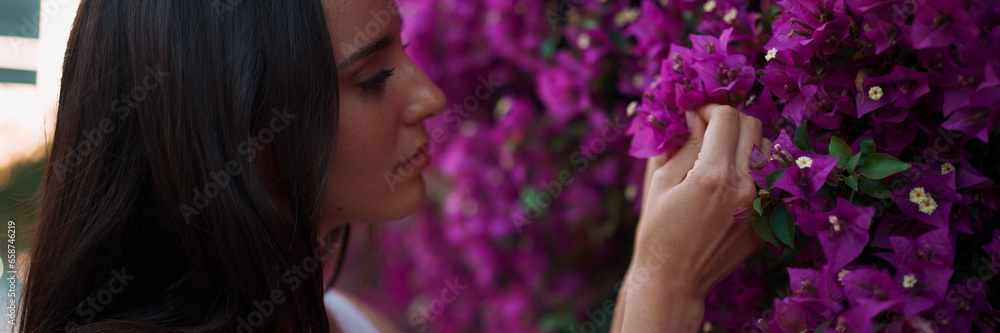 Fototapeta premium Beautiful woman with freckles and dark loose hair wearing white top walks along wall blooming with purple flowers. Cute girl looking at purple flowers