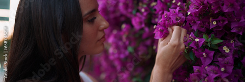 Beautiful woman with freckles and dark loose hair wearing white top walks along wall blooming with purple flowers. Cute girl looking at purple flowers