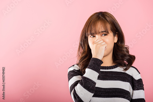 Portrait of an Asian woman, unhappy with a disgusted expression, pinches her nose due to a foul smell. Studio shot isolated on pink, illustrating a healthcare concept.