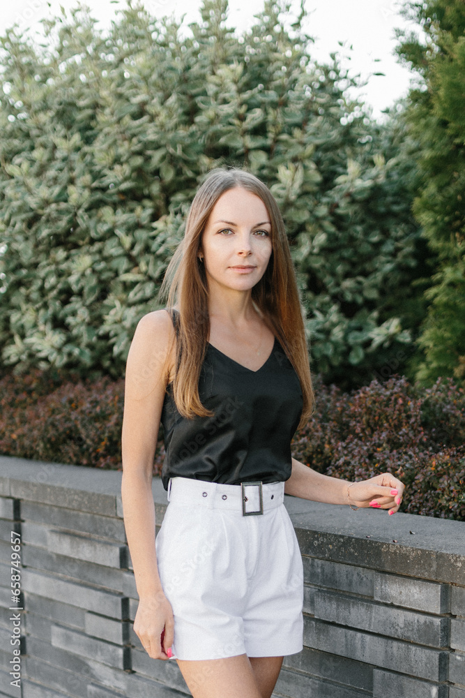 Portrait of a girl with long blond hair, wearing a black top, white shorts, poses against a gray wall. Young woman walking through a park with modern architecture and landscape design Portrait of a girl with long blond hair, wearing a black top, white shorts, poses against a gray wall. Young woman walking through a park with modern architecture and landscape design