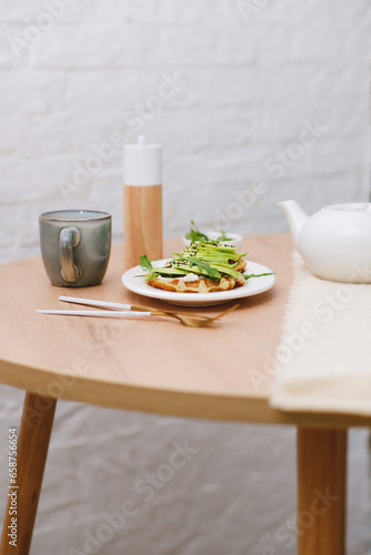 Breakfast on wooden table:zucchini waffles with cottage cheese, avakado and arugula in a white plate, cup of tea. Top view.