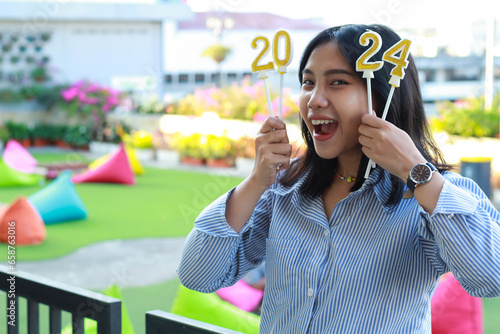 excited young woman asian looking at camera and standing on railing of house yard to celebrate new year eve with holding 2024 number candle, outdoor photo