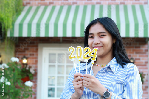 smiling asian young woman looking to camera while standing over brick wall vintage house yard with holding 2024 number candle to celebrating new year eve photo