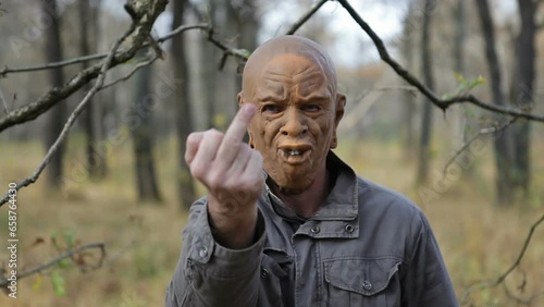 A man in a scary monster mask stands in the forest and shows his middle finger, finger gesturing fuck. A man makes a fuck gesture with his hand against the background of a forest.