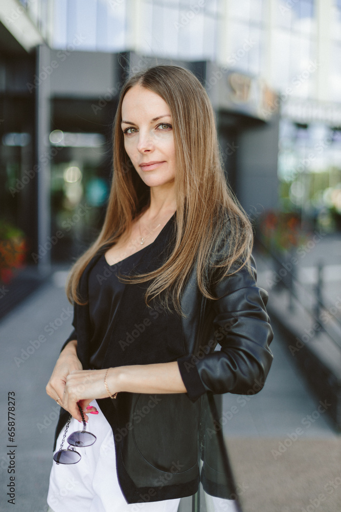 A girl with long blond hair, wearing a black top, black jacket and white shorts, poses against the backdrop of modern architecture. Young woman walking in a park with landscape design