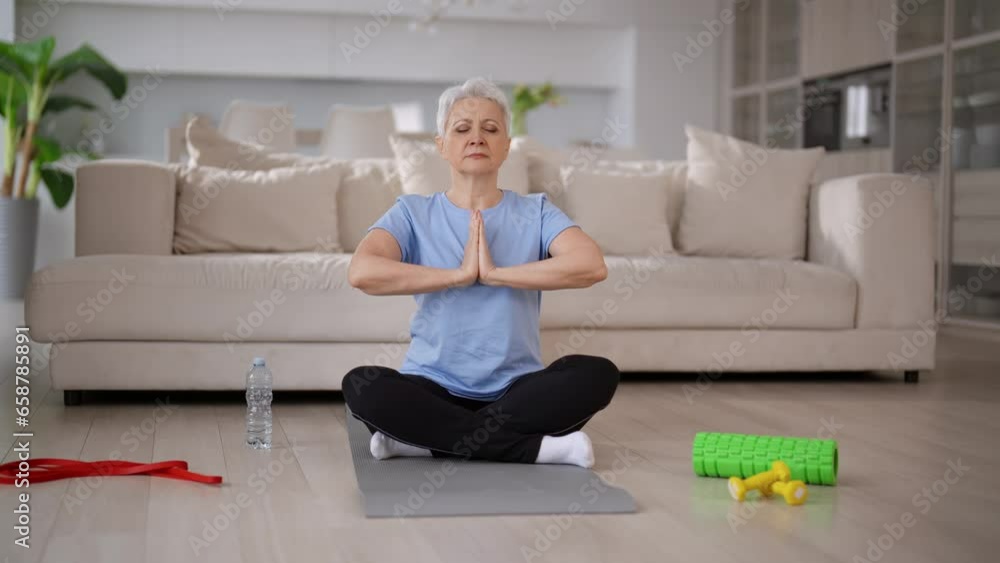 Yoga, sport lifestyle, exercising concept. Relaxed senior woman doing yoga meditation sitting in lotus pose on mat in living room at home. Elderly gray-haired woman making physical exercises. Yoga, sport lifestyle, exercising concept. Relaxed senior woman doing yoga meditation sitting in lotus pose on mat in living room at home. Elderly gray-haired woman making physical exercises.