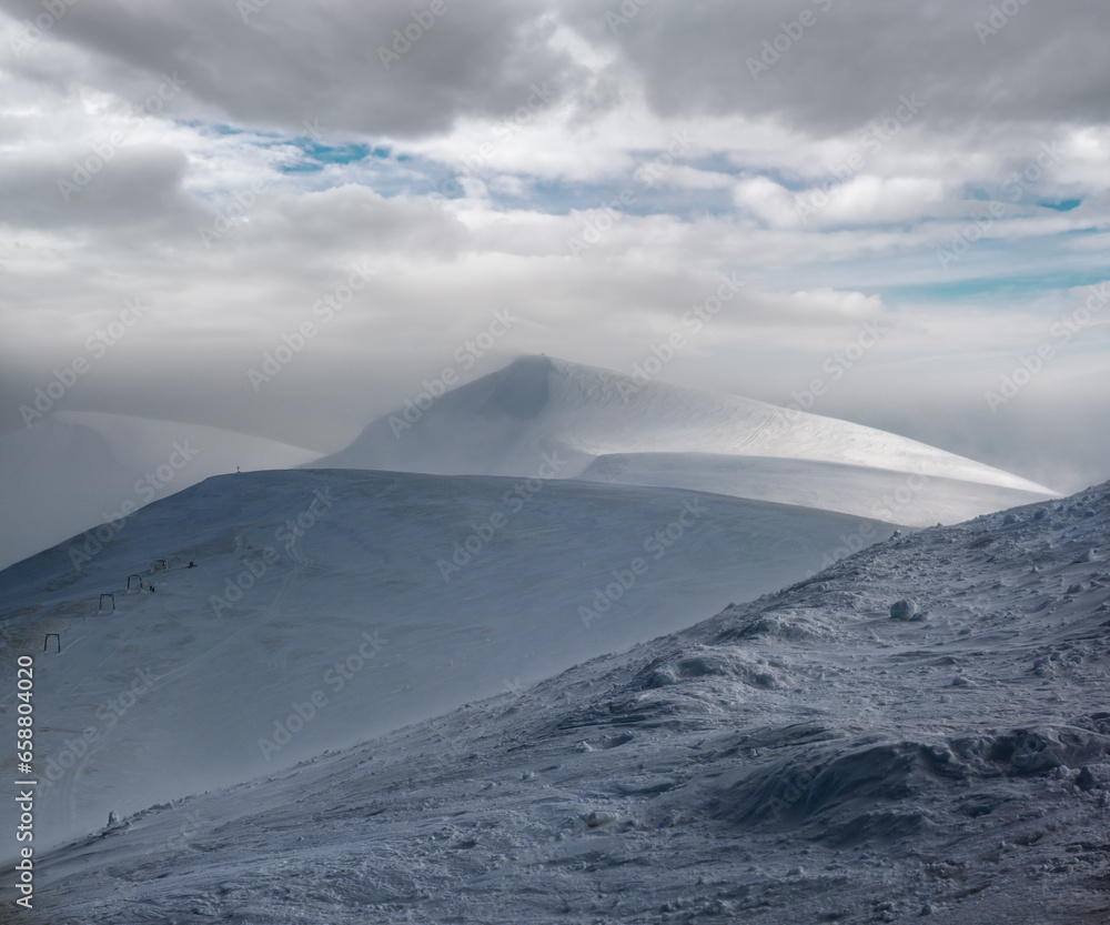 Naklejka premium Snow covered mountain slope in last evening sunlight. Magnificent windy dusk on tops above picturesque alpine ski resort, Dragobrat, Ukraine, Carpathian Mountains.