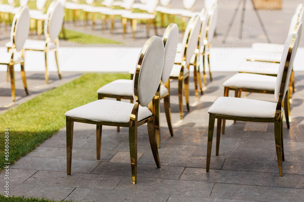 Luxury white-golden chairs on wedding ceremony outdoors. Festive decorations. Empty rows armchairs for guests. Outgoing wedding ceremony in the open air. Decor Studio.