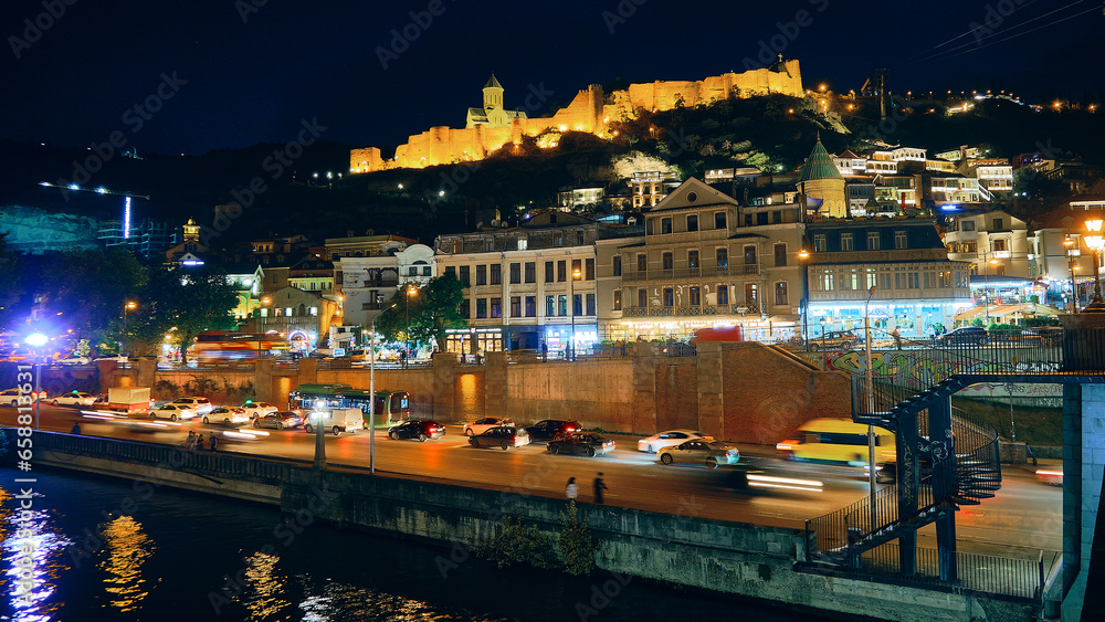 Cars driving on Kura river embankment. Illuminated Narikala fortress at background. Tbilisi night traffic