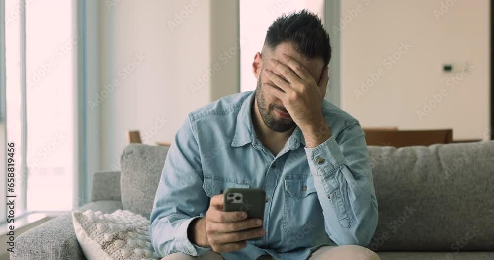 Upset angry Hispanic man sits on sofa hold smartphone staring at screen looks dissatisfied feels irritated due to bad news in message, broken device, wifi connection lost, gadget problem, app crash