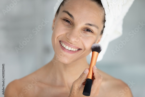 Portrait of smiling millennial female after shower get ready do daily facial make up or beauty procedures in bath  close up of happy young woman use brush put powder on face or cheeks in bathroom