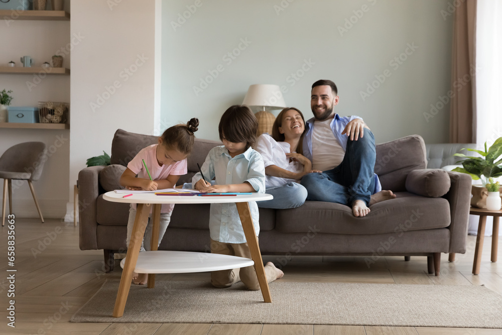 Happy parents relaxing on comfortable sofa at cozy home, watching to little kids drawing at small table, laughing, enjoying weekend, family leisure time, relaxation, good relationship