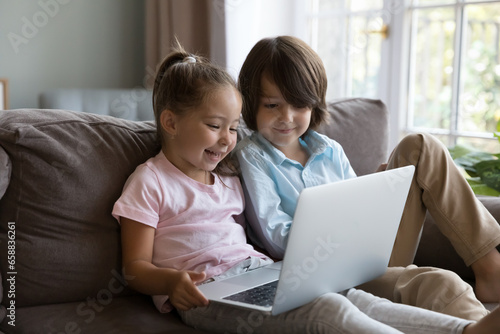 Two happy cute preschool sibling kids talking on video call on laptop, playing online virtual game, watching cartoon movie on Internet, enjoying wireless technology, communication, leisure