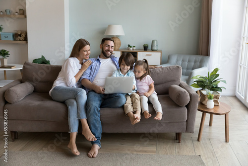 Happy joyful millennial couple and little kids resting on sofa, holding laptop together, watching movie, interactive TV, talking on video call, chatting online, using app for Internet communication