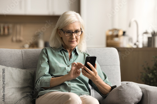 Serious elderly blonde woman in eyewear typing on cellphone at home, using medical application, healthcare service on smartphone, wireless Internet connection, sitting on home sofa © fizkes