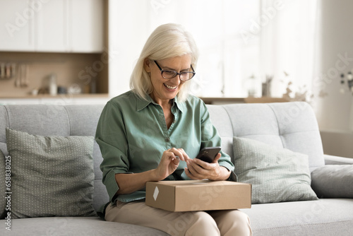 Cheerful old retired consumer woman getting purchase from Internet store, holding paper box on lap, using smartphone, giving positive review, feedback to delivery logistic service, Internet store © fizkes