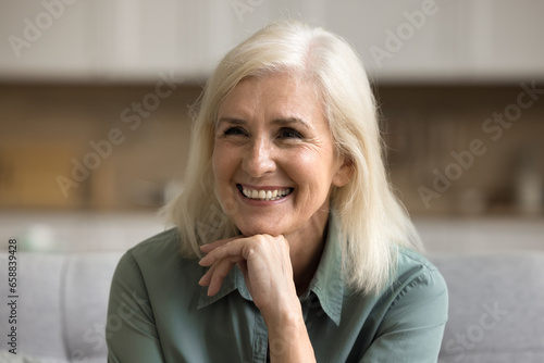 Happy blonde elderly woman having fun at home, leaning chin on hand, looking away with toothy smile, laughing, enjoying retirement, positive emotions. Close up casual portrait