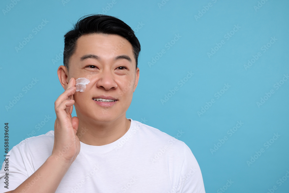Handsome man applying cream onto his face on light blue background. Space for text Handsome man applying cream onto his face on light blue background. Space for text