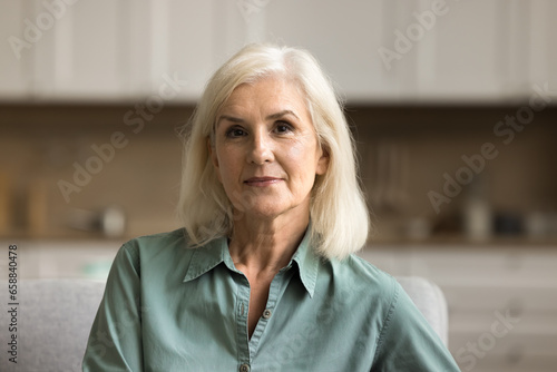 Pretty blonde old mature woman looking at camera with serious positive face expression, posing indoors in modern home apartment interior. Video conference call head shot portrait photo