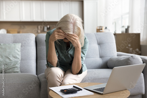 Frustrated tired blonde older woman touching head at calculator and laptop at home, covering face with hand, getting stress, problems, bad news, suffering from depression