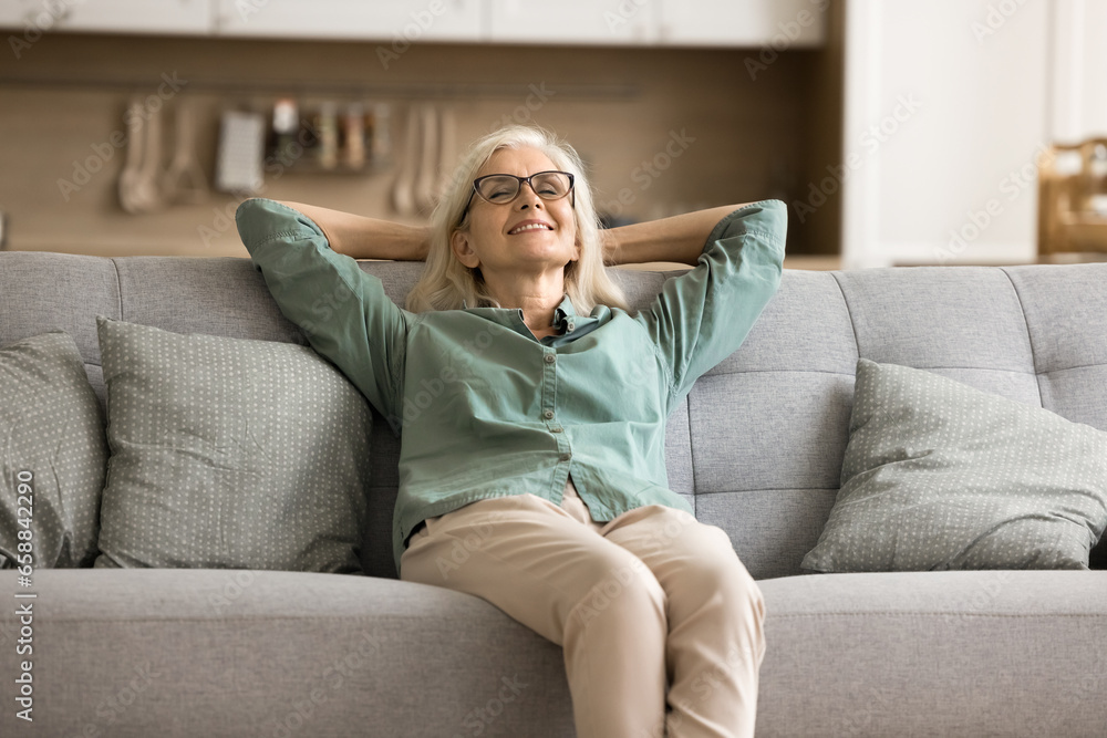 Fototapeta premium Cheerful blonde elderly woman in glasses enjoying comfort at cozy home, resting on comfortable couch, leaning on soft back with closed eyes, taking deep breath, dreaming, smiling