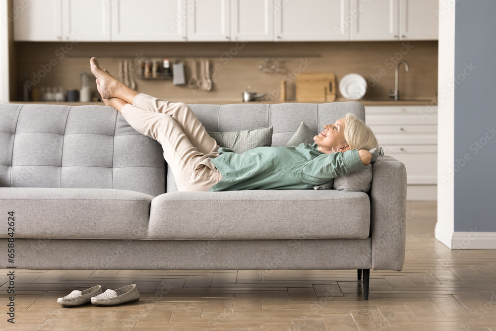 Cheerful sleepy blonde older lady lying on couch, leaning legs and feet on soft back, enjoying leisure, lazy break in modern home interior, breathing fresh cool pure air with closed eyes - obrazy, fototapety, plakaty 