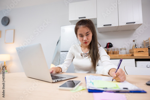 writing note or journal on note book. Reading email and working. Asian thai young woman sitting working on laptop computer. Thai female using laptop and smiling at home.