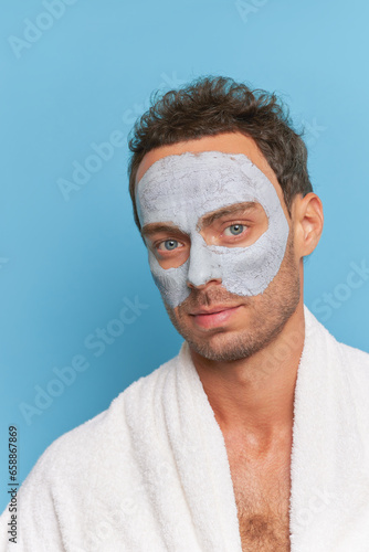Portrait of handsome model guy with clay facial mask and towel on shoulders, cosmetic treatments for men concept, copy space